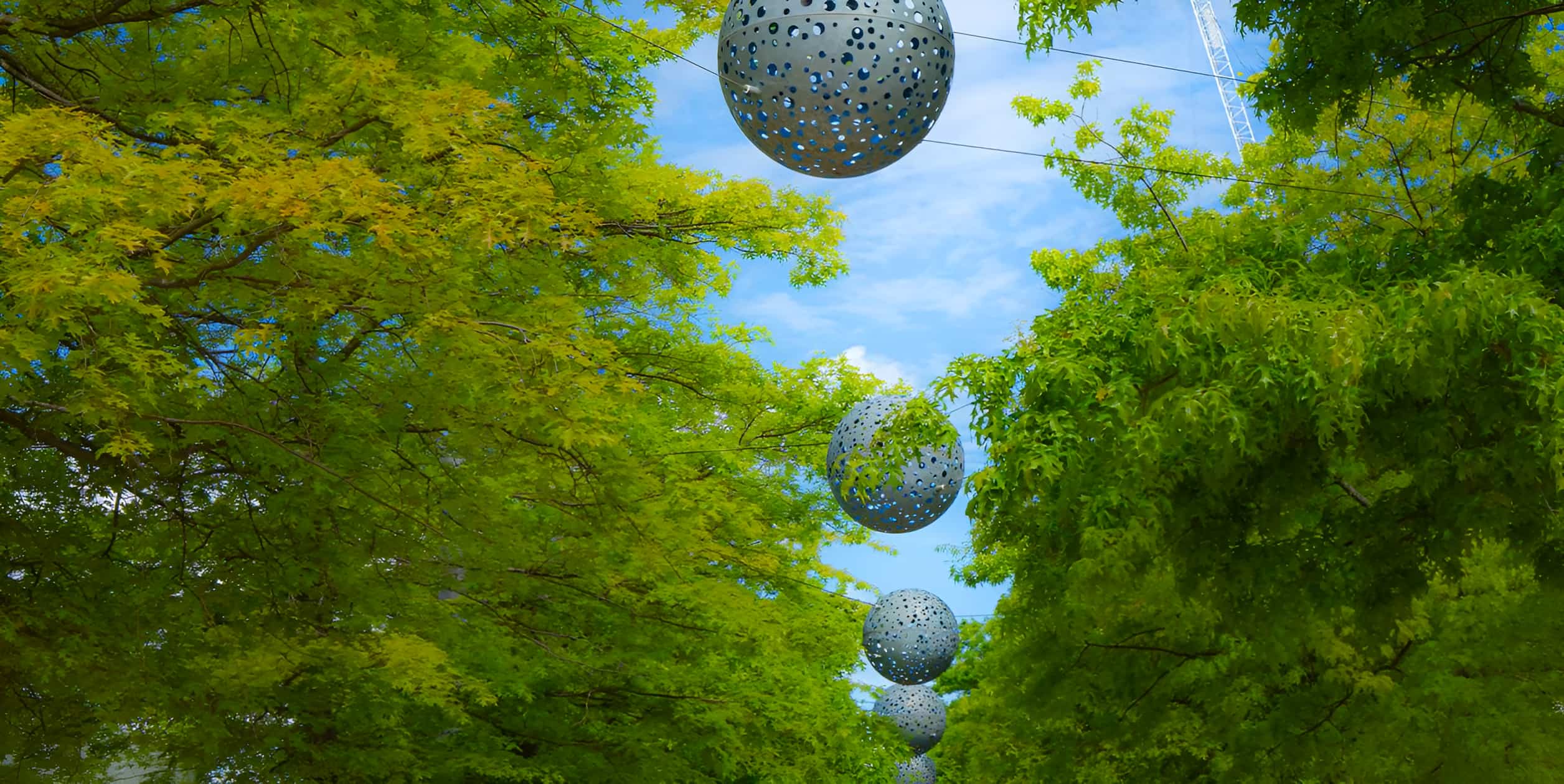 Perforated metal sphere sculptures suspended between lush green trees against a blue sky in a landscaped avenue