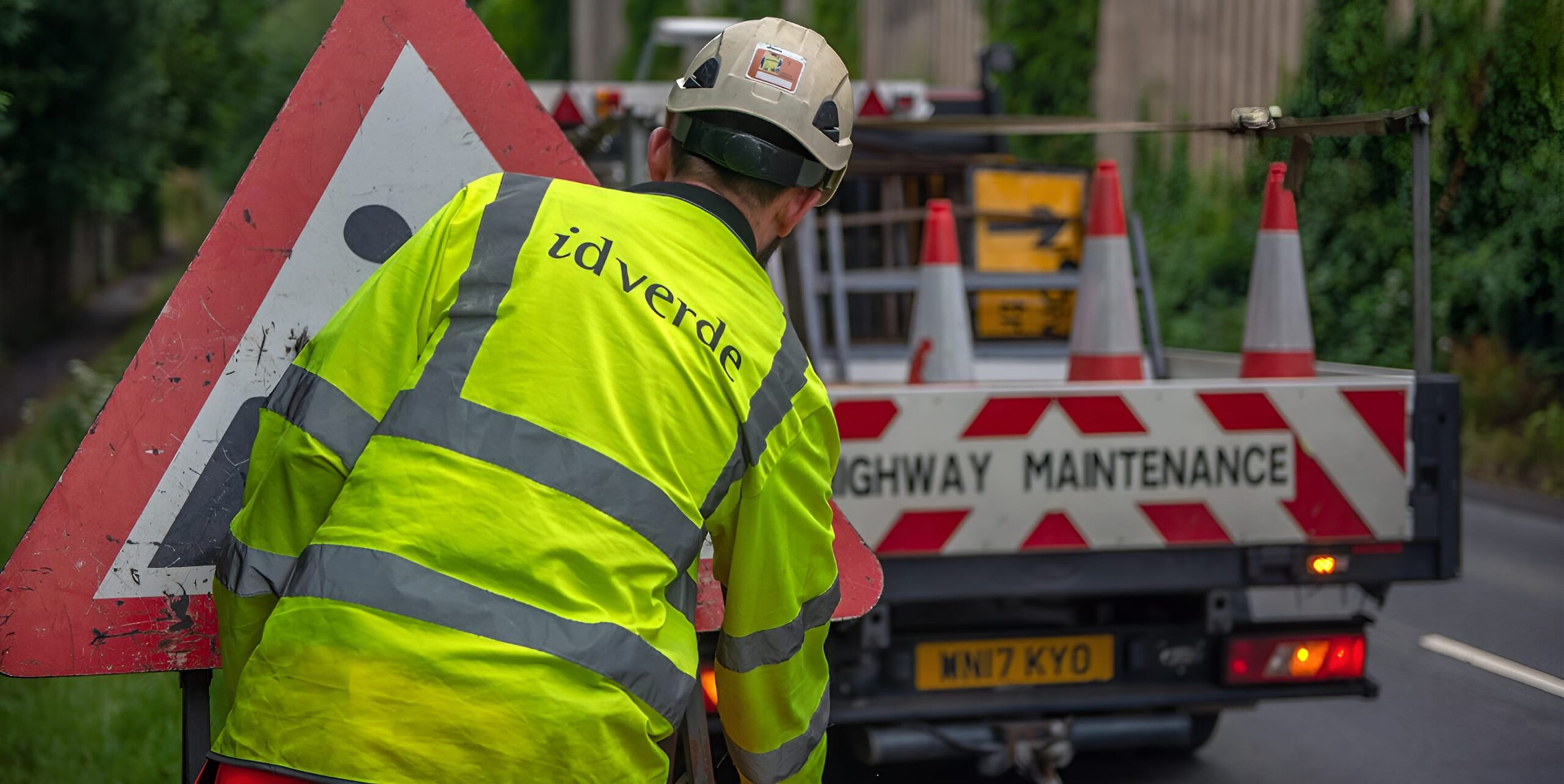 idverde highway maintenance worker in hi-vis jacket placing road warning sign beside a maintenance truck with traffic cones