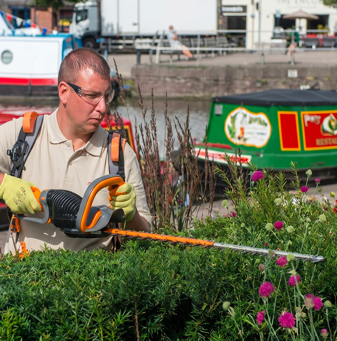 An idverde operative trimming a low hedge with a battery-powered hedge cutter on a canalside planting scheme, with narrowboats moored in the background.
