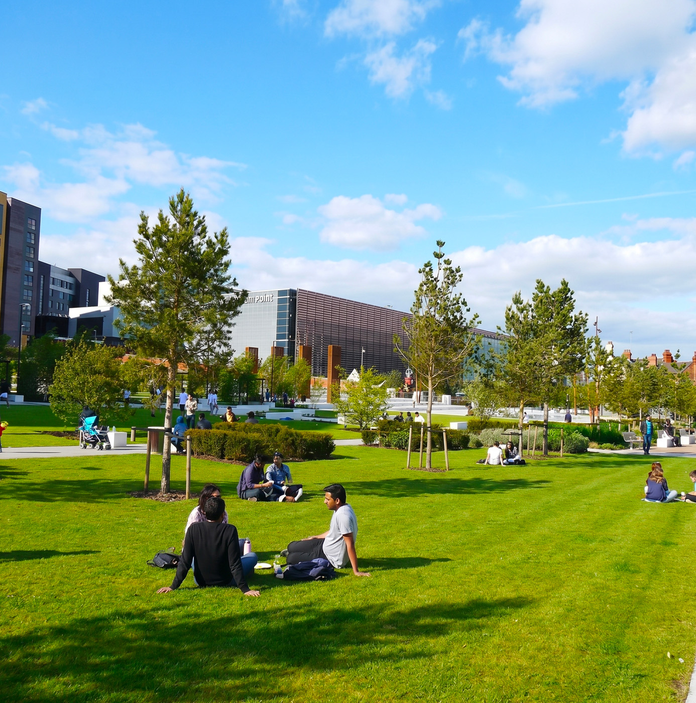 People relaxing on a well-maintained lawn in a contemporary urban park with young trees, planted borders, and a modern building in the background.
