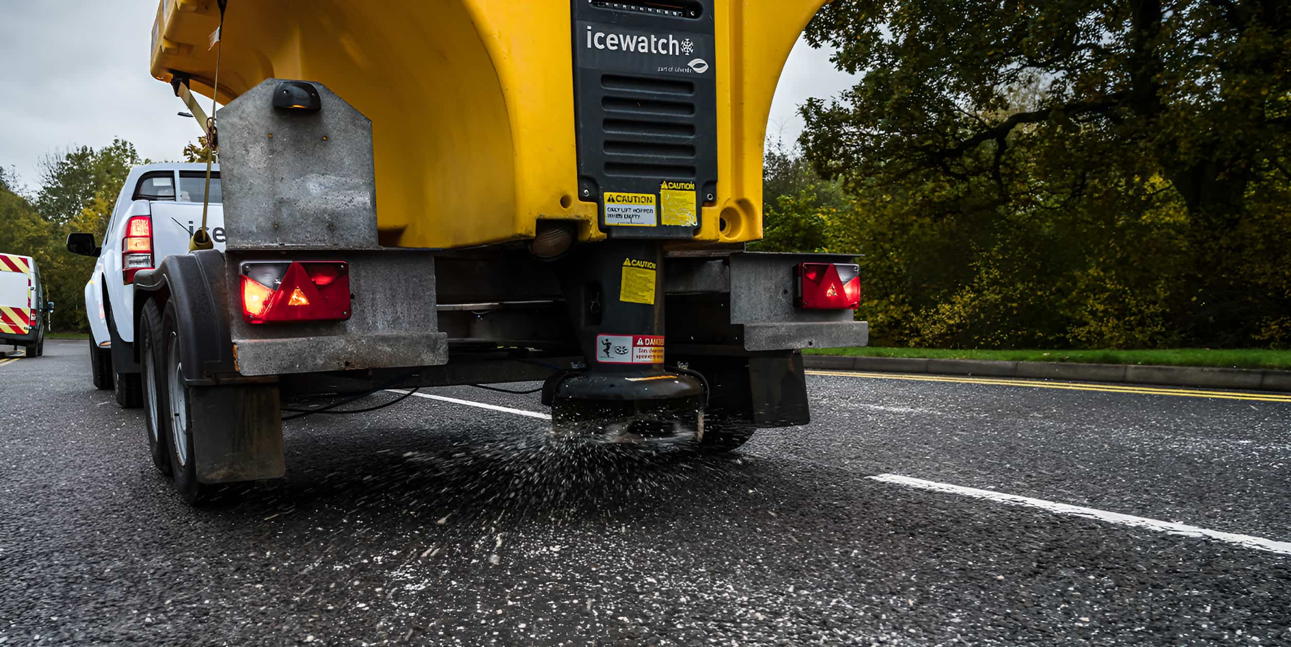 Icewatch gritter trailer spreading salt on a wet road, towed by a white pickup truck