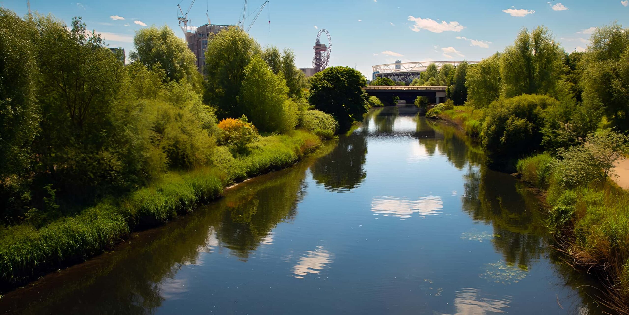 River Lea flowing through Queen Elizabeth Olympic Park with London Stadium and ArcelorMittal Orbit visible beyond lush green trees