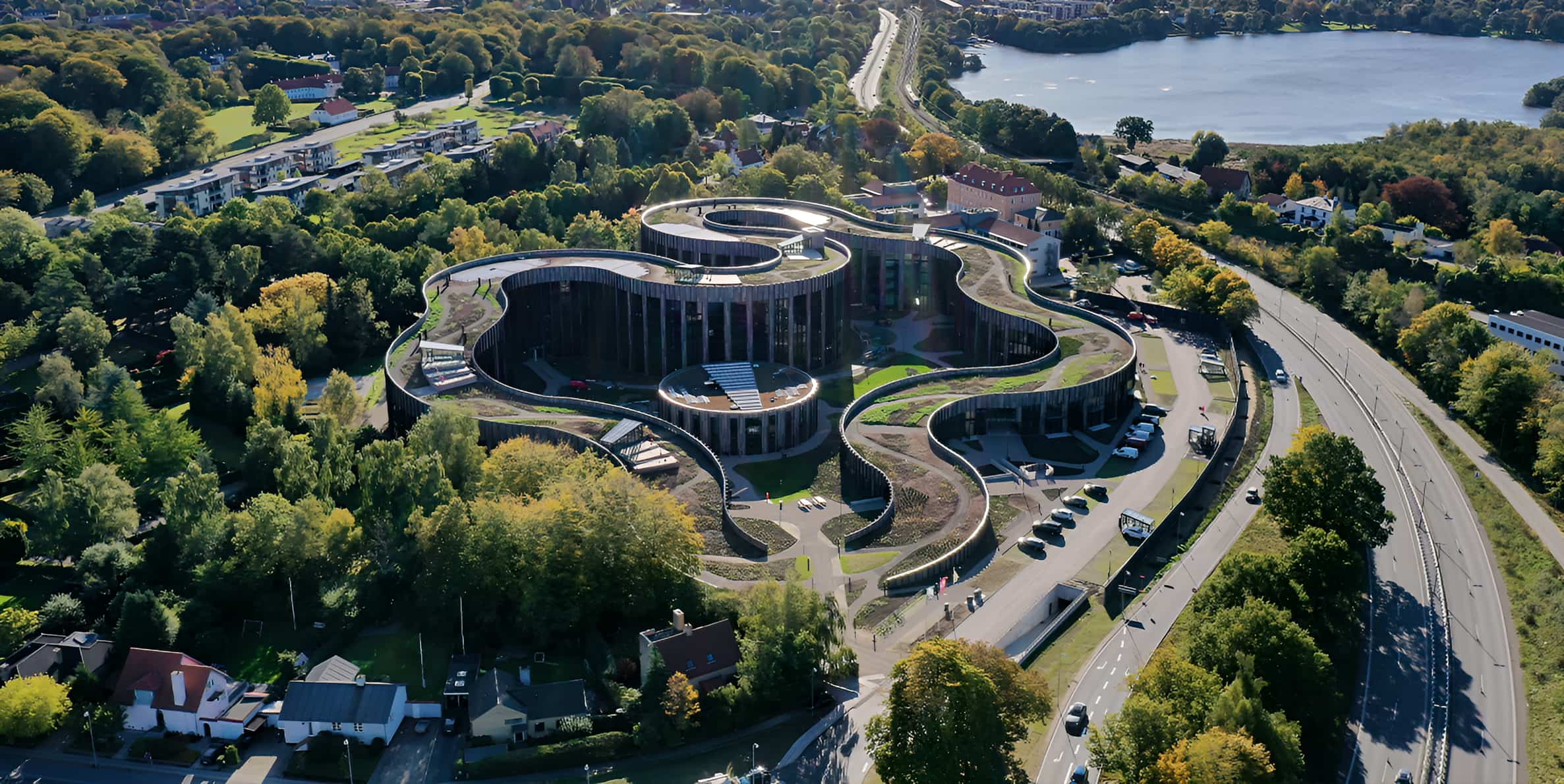 Aerial view of a flower-shaped building with green rooftop gardens surrounded by trees and a lake in Scandinavia