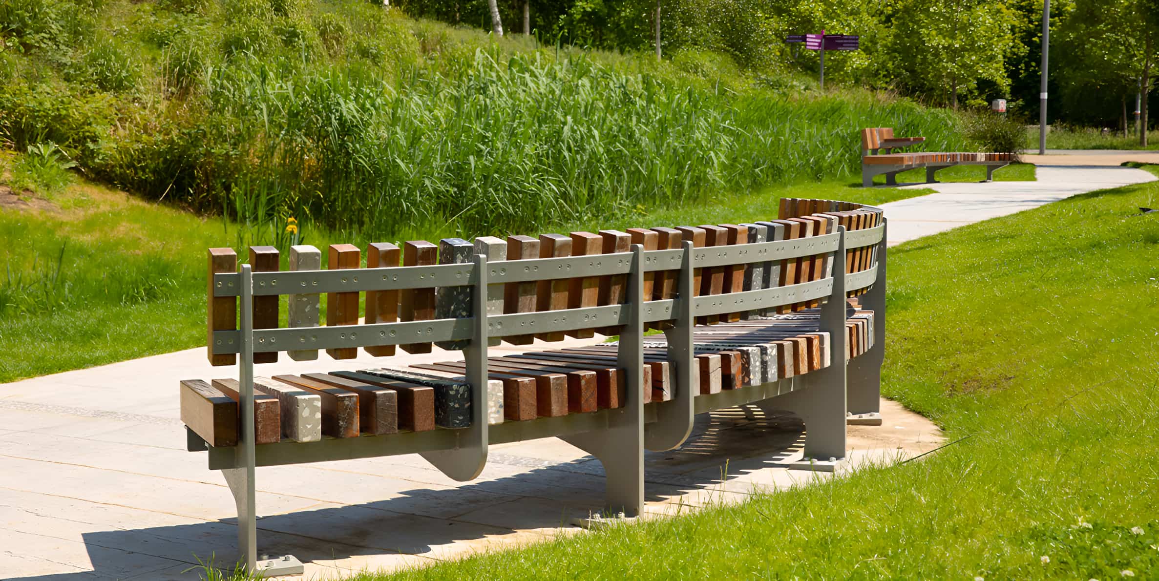 Curved park benches with mixed wood and stone slats on metal frames beside a paved path in a green landscaped park