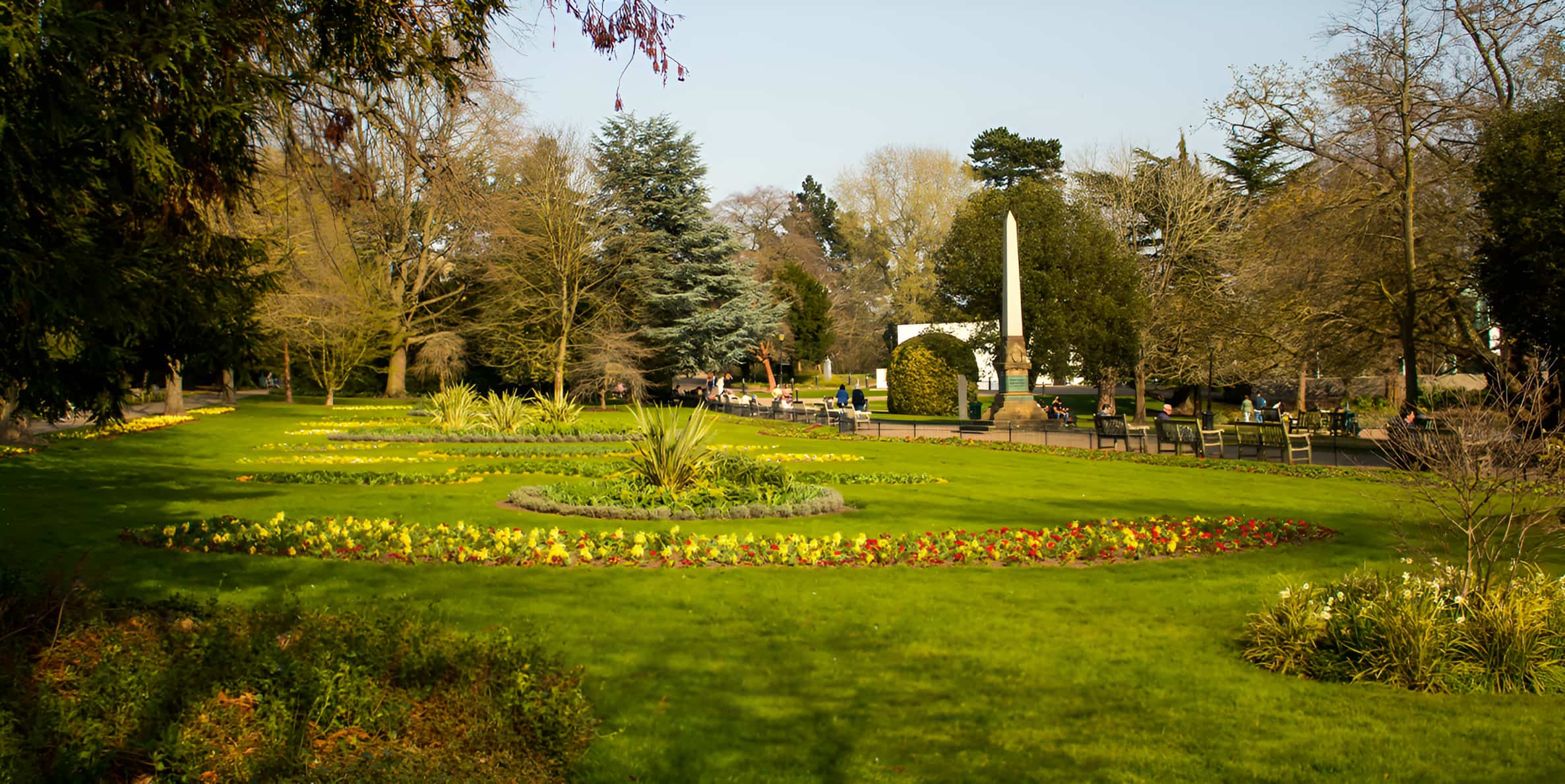 Formal park garden with colourful flower beds, obelisk monument, manicured lawns and mature trees on a sunny spring day