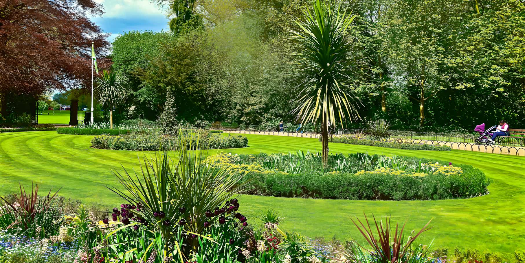 Manicured public park with circular flower beds, cordyline palms, striped lawn, and mature trees on a sunny day