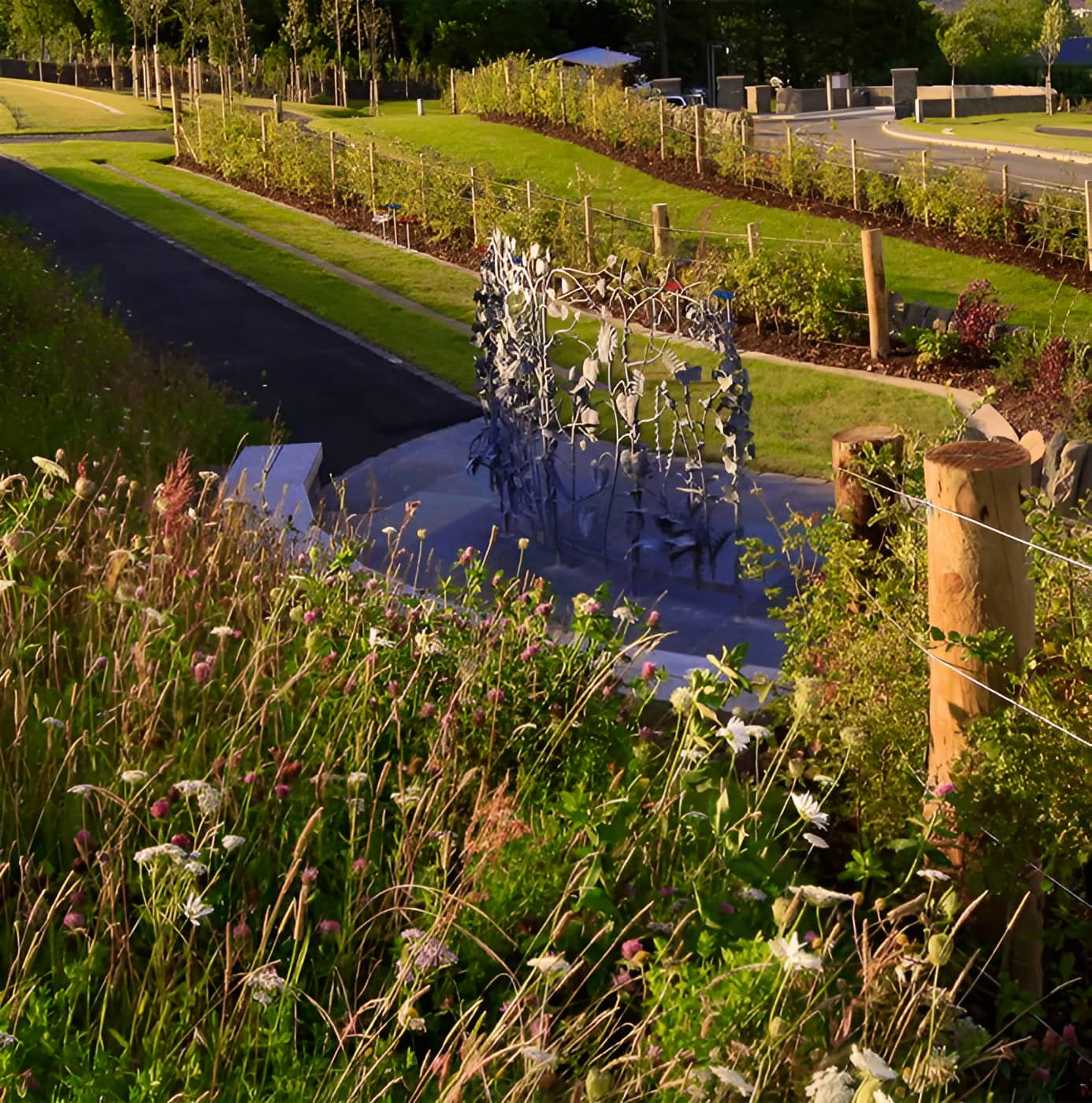 Metal botanical sculpture beside a water feature, surrounded by wildflower meadow and landscaped grounds with young trees
