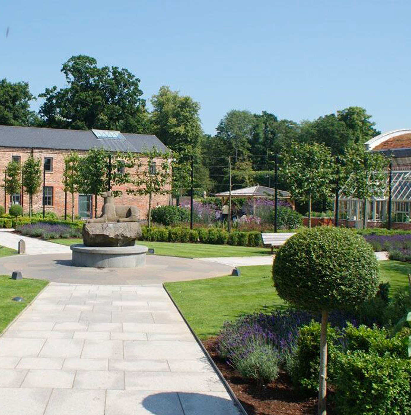 A formal garden courtyard with stone paving, a central sculpture on a circular plinth, ball-shaped topiary, lavender borders, and a restored Victorian glasshouse.