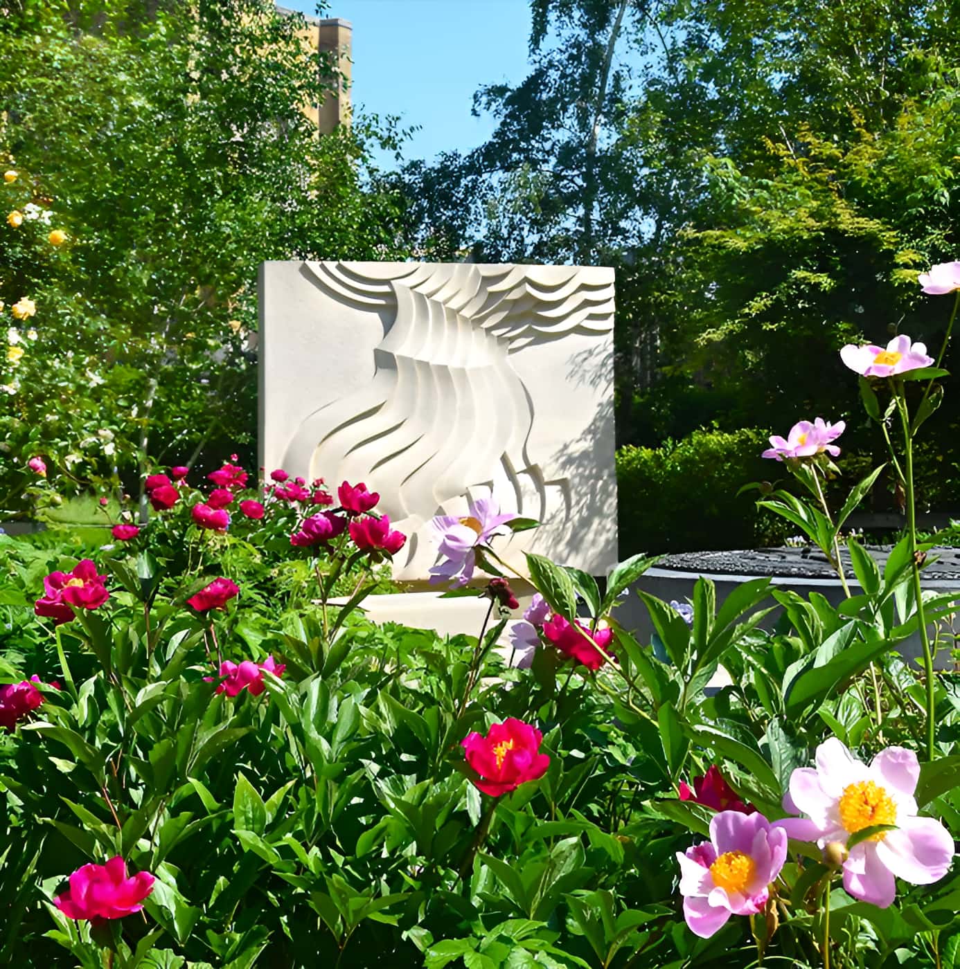 White abstract stone sculpture with wave carvings surrounded by red peonies and pink flowers in a landscaped garden