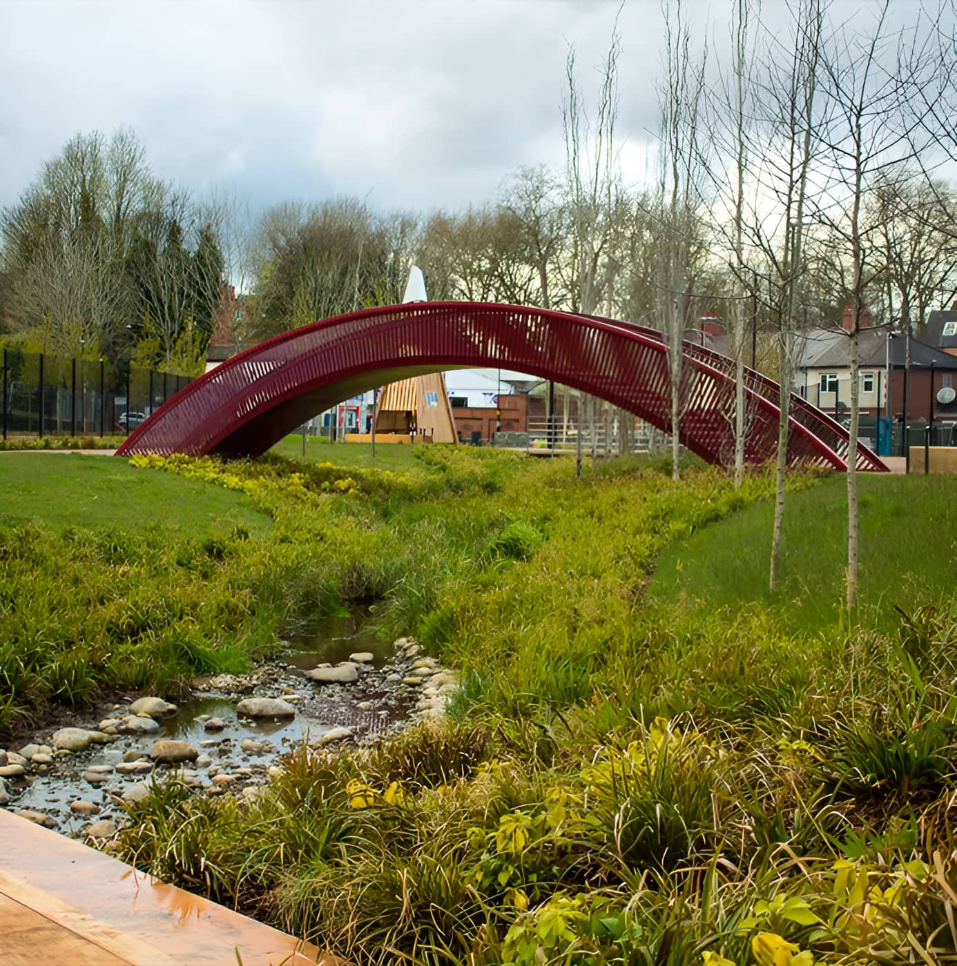 Red arched footbridge over a rocky stream in a landscaped urban park with wildflower grass and bare trees