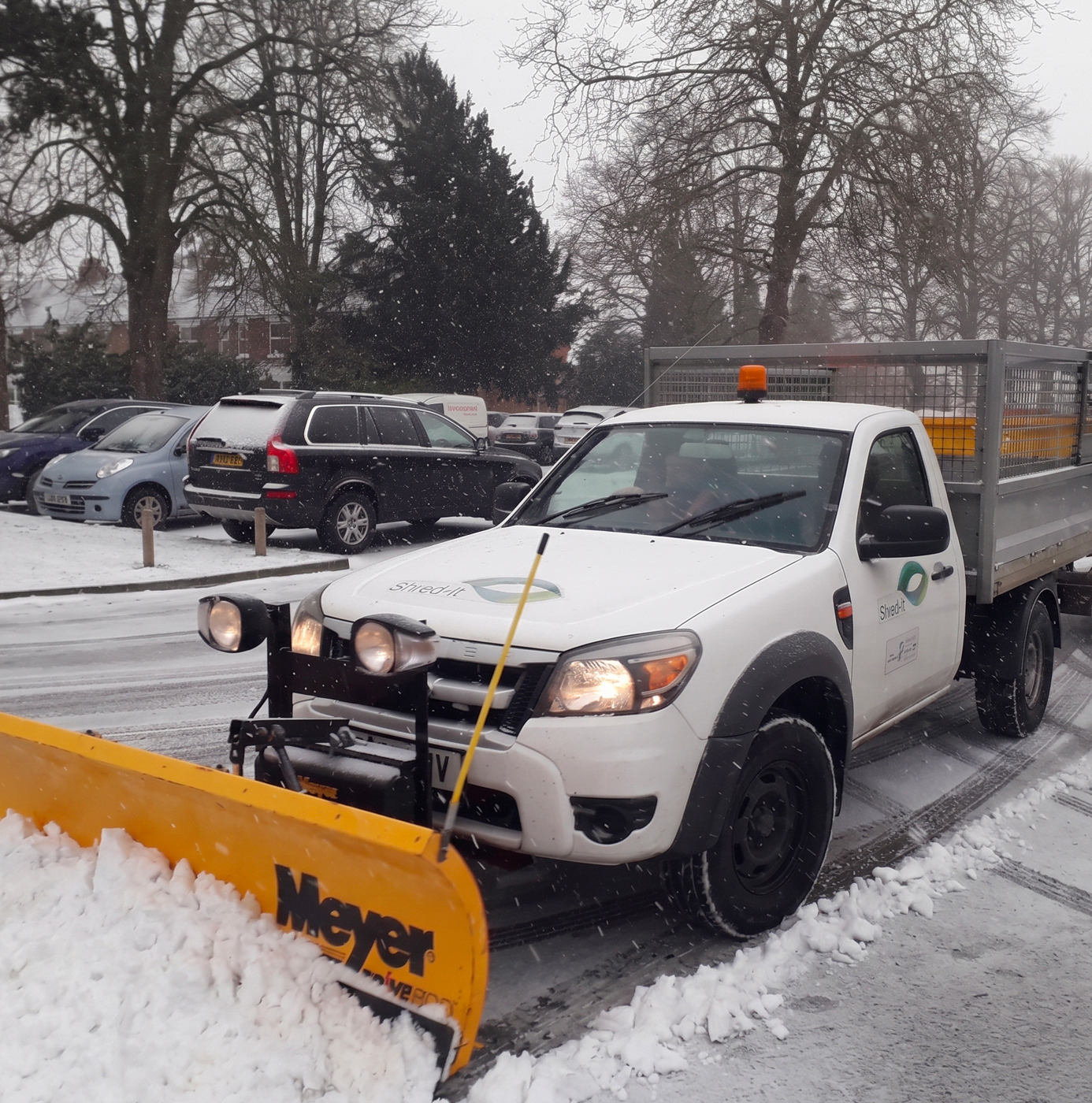 A white pick-up truck fitted with a yellow Meyer snow plough clearing a snow-covered car park during heavy snowfall
