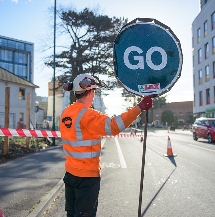 idverde worker in orange hi-vis holding a GO lollipop sign on a road during construction works