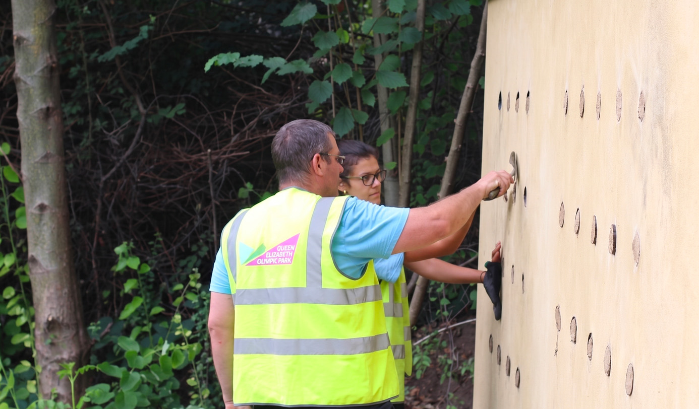 Two operatives in high-visibility vests installing cork-plugged bee bricks into a large insect hotel structure in a woodland edge setting
