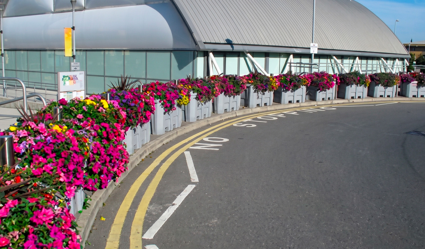 A long row of grey container planters overflowing with pink, red, and yellow summer flowers along the entrance road of a large public venue