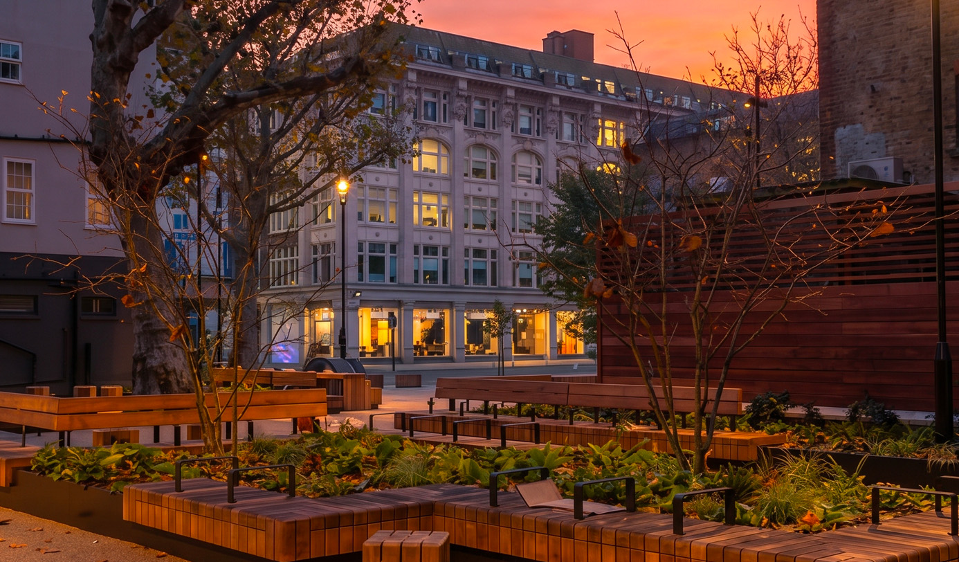 A newly landscaped urban courtyard at dusk featuring timber bench seating, integrated planters with shade-tolerant planting, and mature trees lit by street lamps.