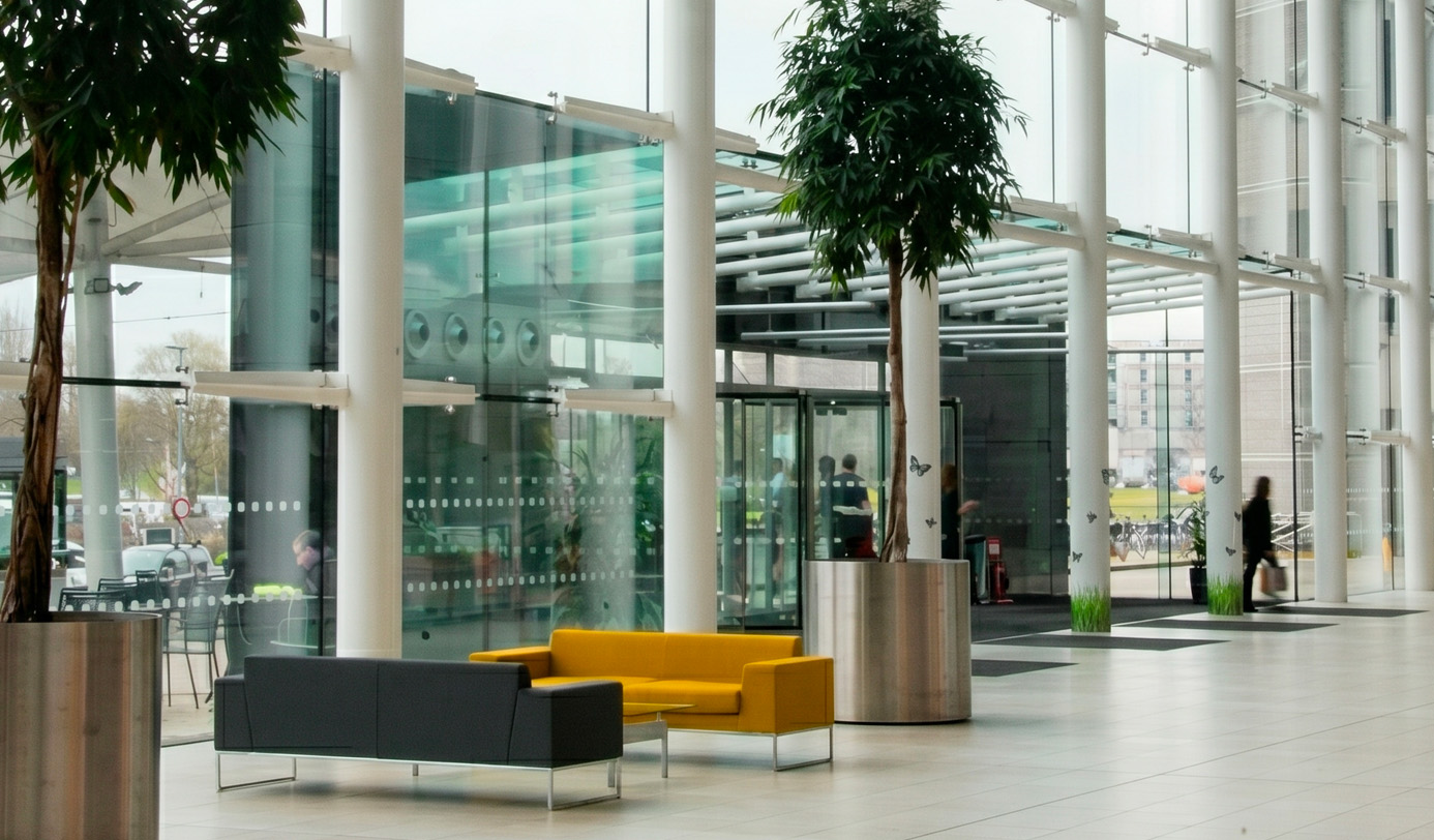 Tall indoor trees in brushed steel planters in a high-ceiling glazed entrance lobby of a modern public building.