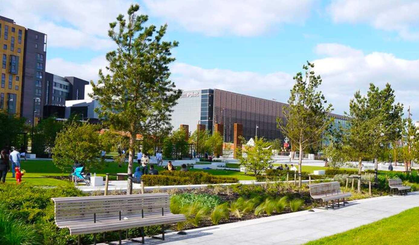 People relaxing in a designed urban park with specimen pine trees, mixed planted borders, timber benches, and a contemporary public building in the background.
