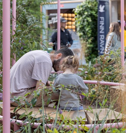 Adult bending down to interact with a young child on a wooden deck surrounded by lush garden plants and pink metal structures