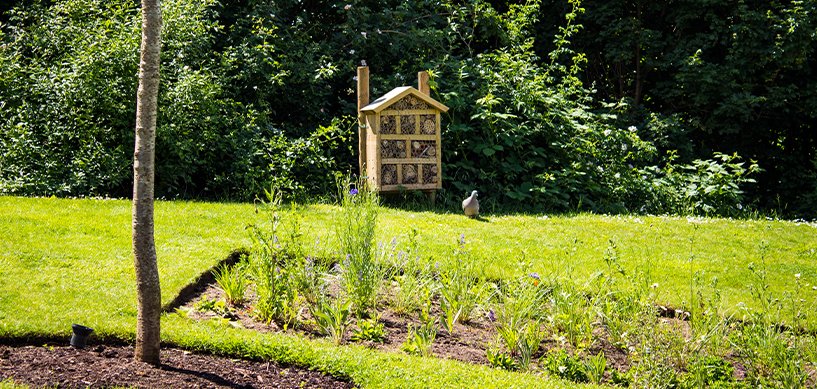 Wooden insect hotel on a post in a landscaped garden with wildflower bed, green lawn and trees