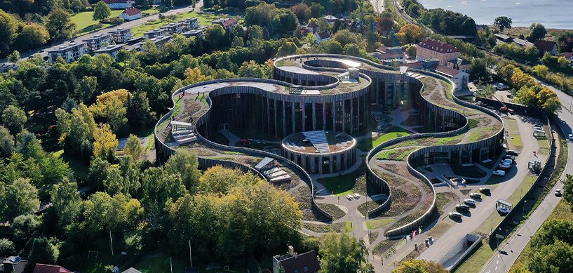 Aerial view of a curved green-roofed building complex surrounded by lush trees near a coastline