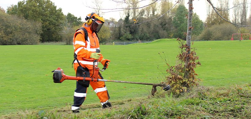 idverde groundsworker in orange hi-vis PPE using a grass strimmer to trim edges around a young tree in a park