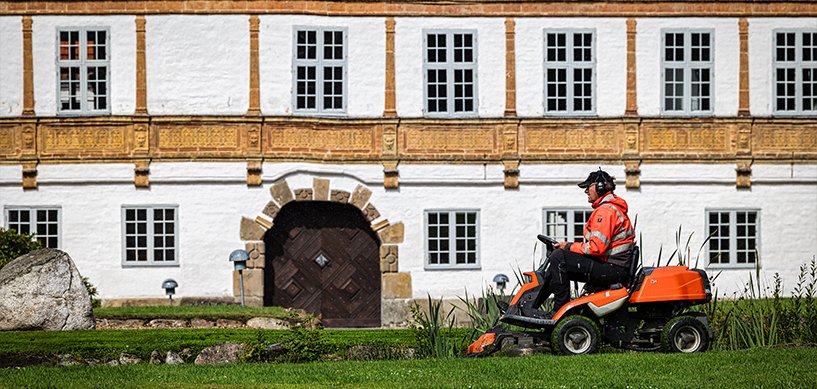 Grounds maintenance worker on orange ride-on mower tending lawn outside a historic white manor building