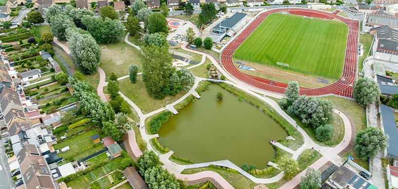 Aerial view of urban park with pond, winding paths, mature trees, and adjacent red athletics track in a French town.