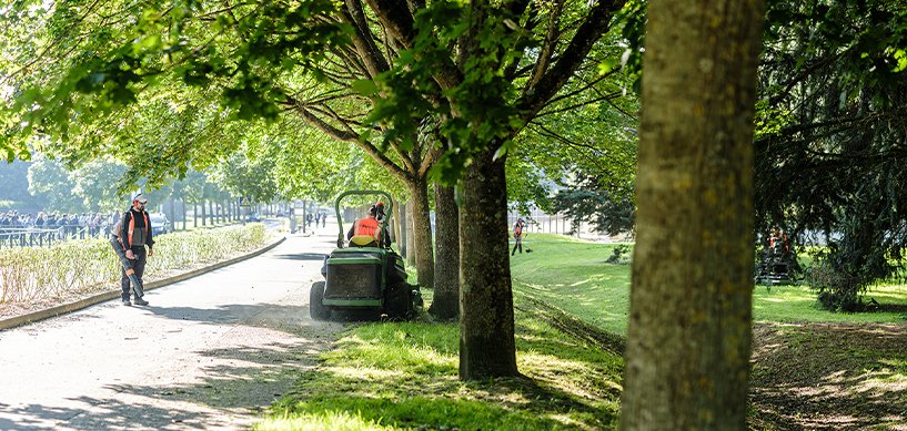 idverde grounds crew maintaining a tree-lined park path with a ride-on mower and workers in hi-vis vests