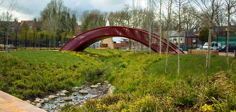 Red arched pedestrian bridge over a rocky stream in a landscaped urban park with green meadow planting