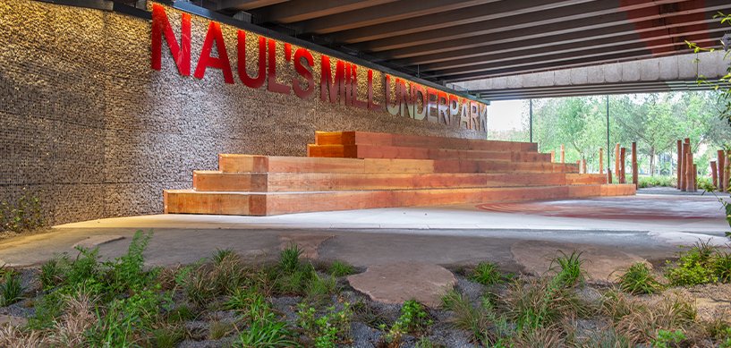 Naul's Mill Underpark featuring timber seating steps, gabion wall signage, and landscaped greenery beneath an overpass