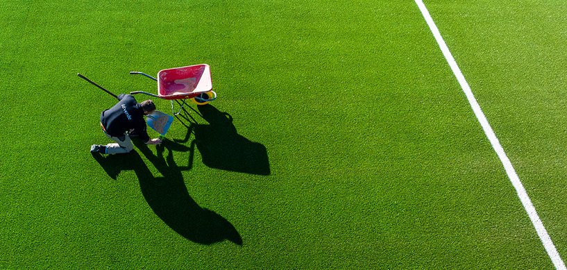 idverde worker kneeling on artificial turf pitch with wheelbarrow, maintaining sports field markings from aerial view