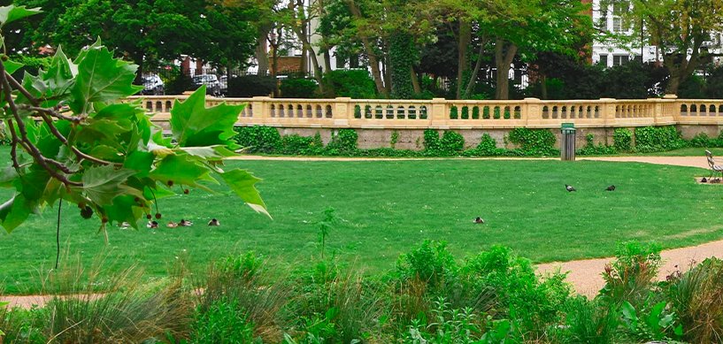 Manicured green lawn with ornate stone balustrade wall, ivy hedging, and birds resting on grass in a formal park setting