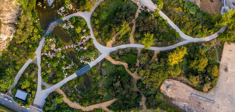 Aerial view of a landscaped park with winding paths, lush greenery, a pond, and stepping stones.