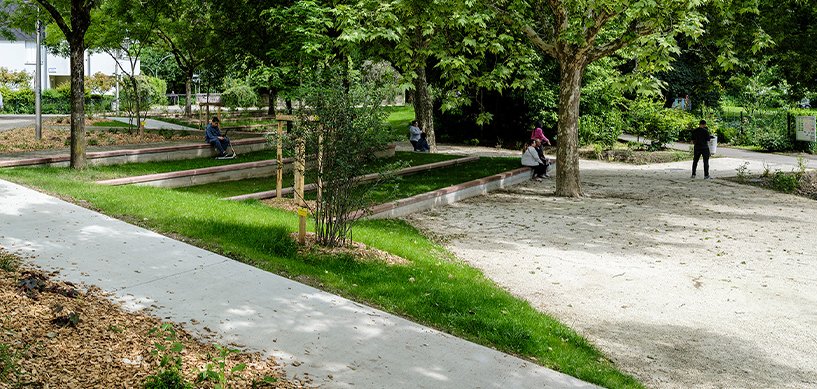 Urban park with manicured lawns, sandy pétanque courts, young trees, and visitors relaxing on benches