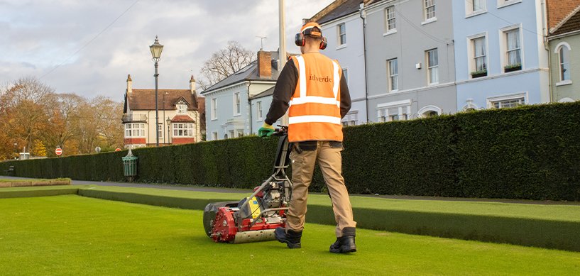 idverde groundskeeper in hi-vis vest mowing a pristine green lawn beside a tall hedge in an urban park
