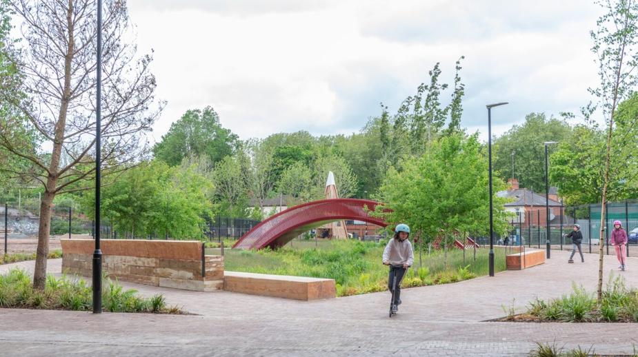 Child riding a scooter through a modern urban park featuring a striking red arch bridge and lush landscaping