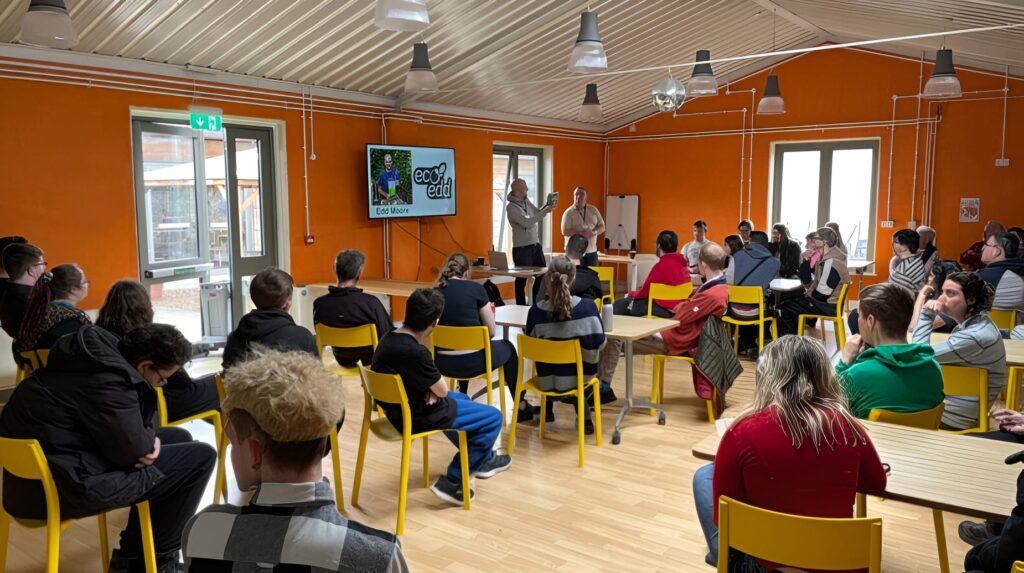Eco Edd presenter Edd Moore speaks to a group seated on yellow chairs in an orange-walled room