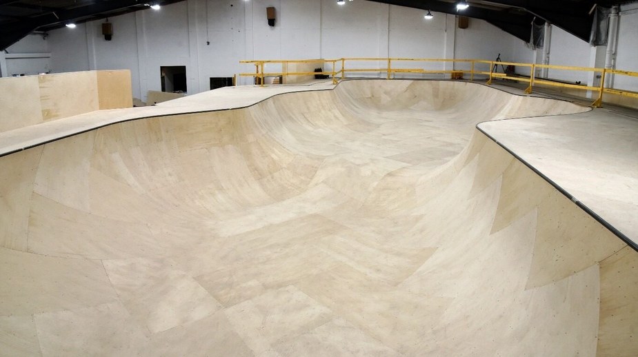 Large indoor wooden skate park bowl with curved walls inside a warehouse facility with yellow safety railings