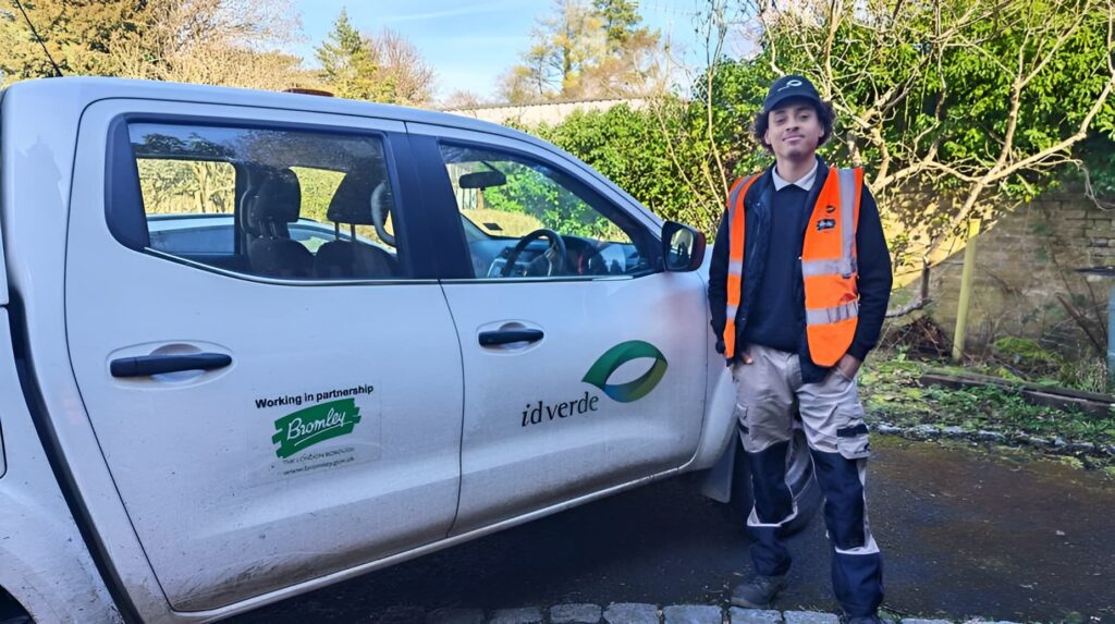 idverde operative in orange hi-vis vest standing beside a white idverde-Bromley partnership pickup truck outdoors.