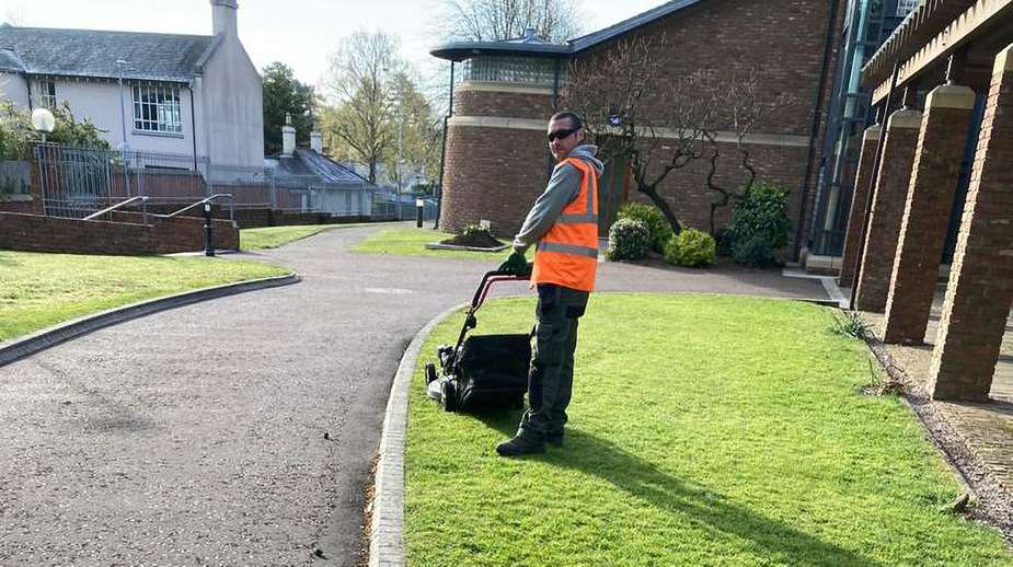 idverde grounds maintenance operative in hi-vis vest mowing a neat lawn beside a brick building and curved driveway