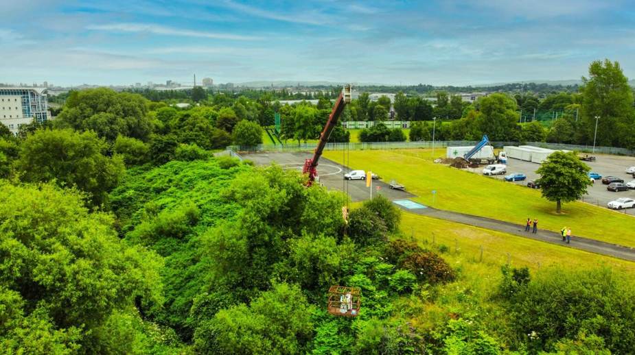Aerial view of crane lifting equipment through dense tree canopy at a managed greenspace site with open grounds and parked vehicles