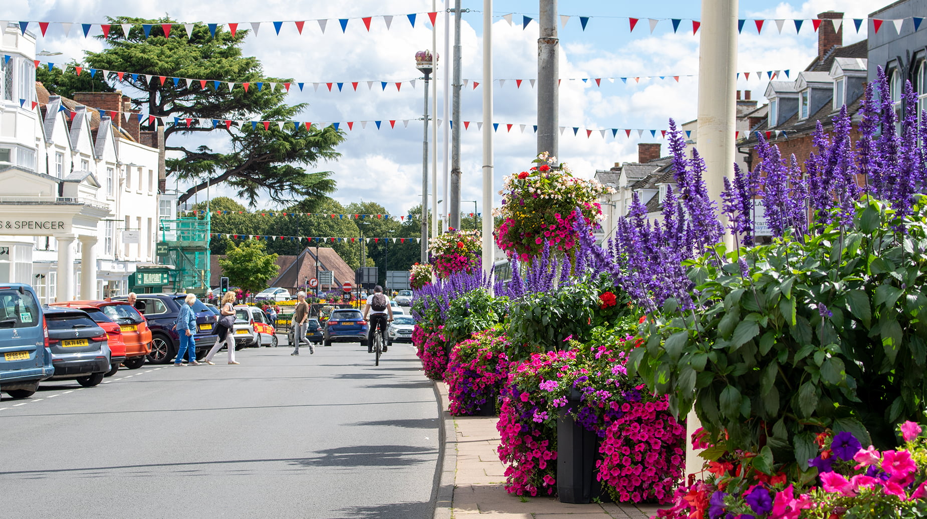 Vibrant floral displays with purple salvia and pink petunias lining a busy UK high street decorated with bunting