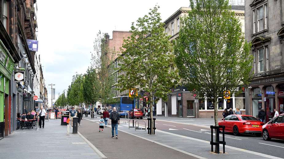 Tree-lined urban street with young deciduous trees, wide pedestrian pavements, and mixed-use retail buildings in a UK city centre