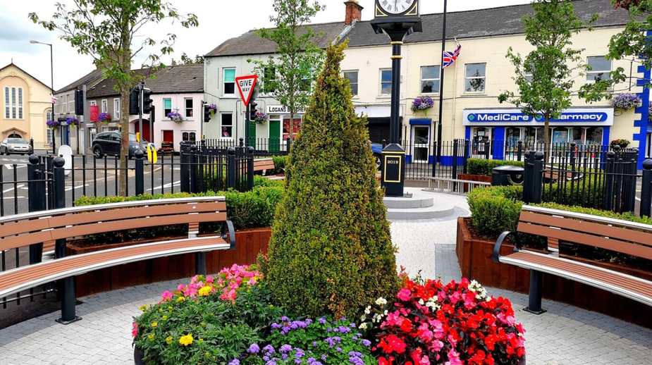 Landscaped town centre public space with colourful flower beds, topiary, curved wooden benches, and a clock tower in Northern Ireland