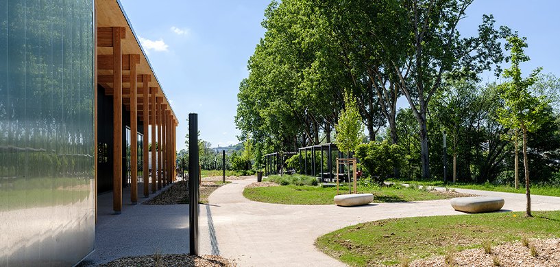 Modern timber-clad building beside a curved pathway with concrete benches, lush trees and landscaped gardens on a sunny day