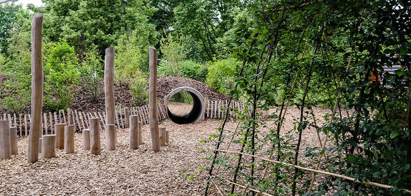 Natural play area with wooden log posts, wood chip ground cover, and a concrete tunnel set into a mound surrounded by trees