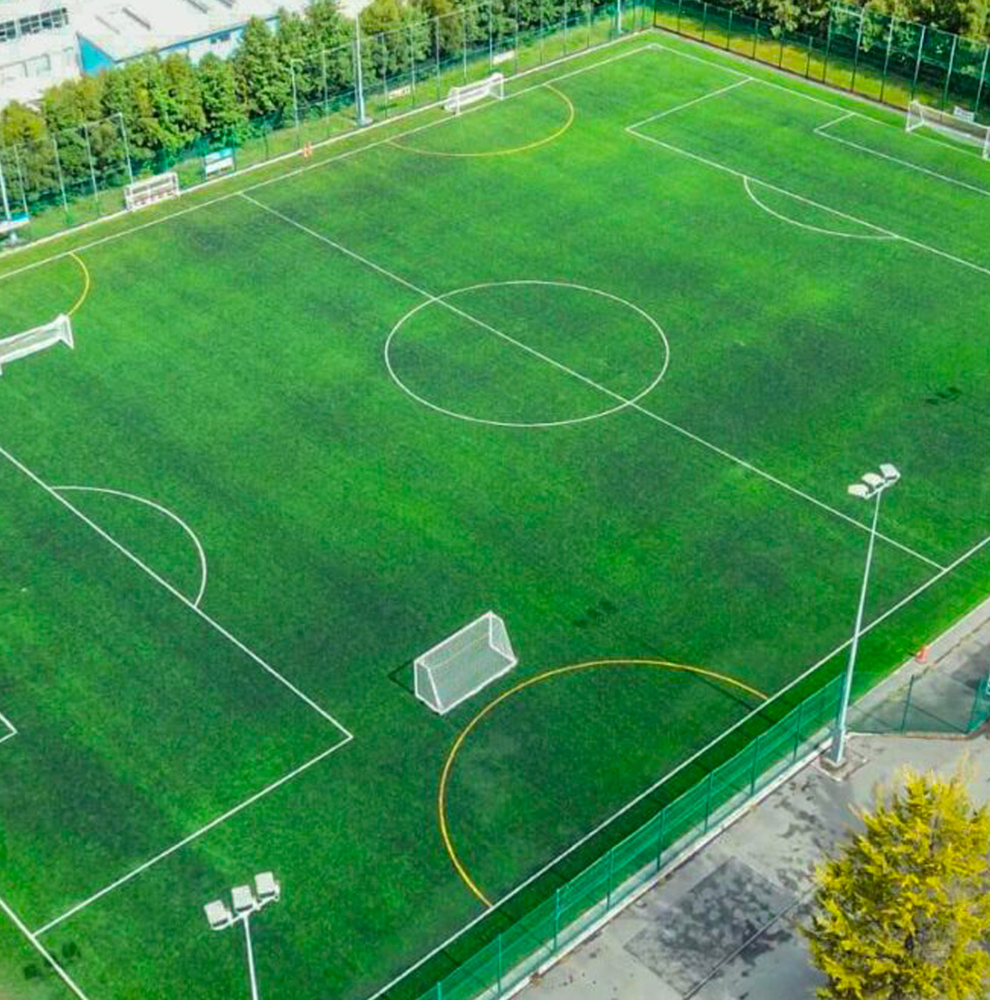 Aerial view of a full-size artificial grass football pitch with white and yellow line markings, perimeter fencing, and floodlights.