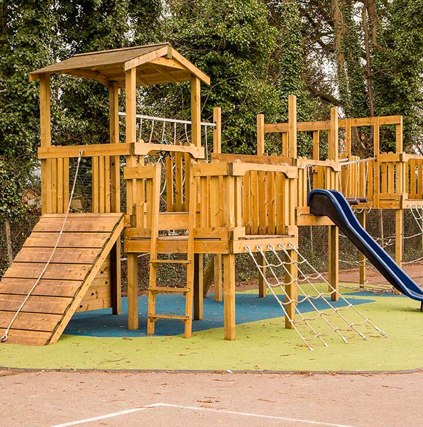 A multi-level timber play structure with slide, rope net, climbing ramp, and elevated walkways on a rubberised safety surface.
