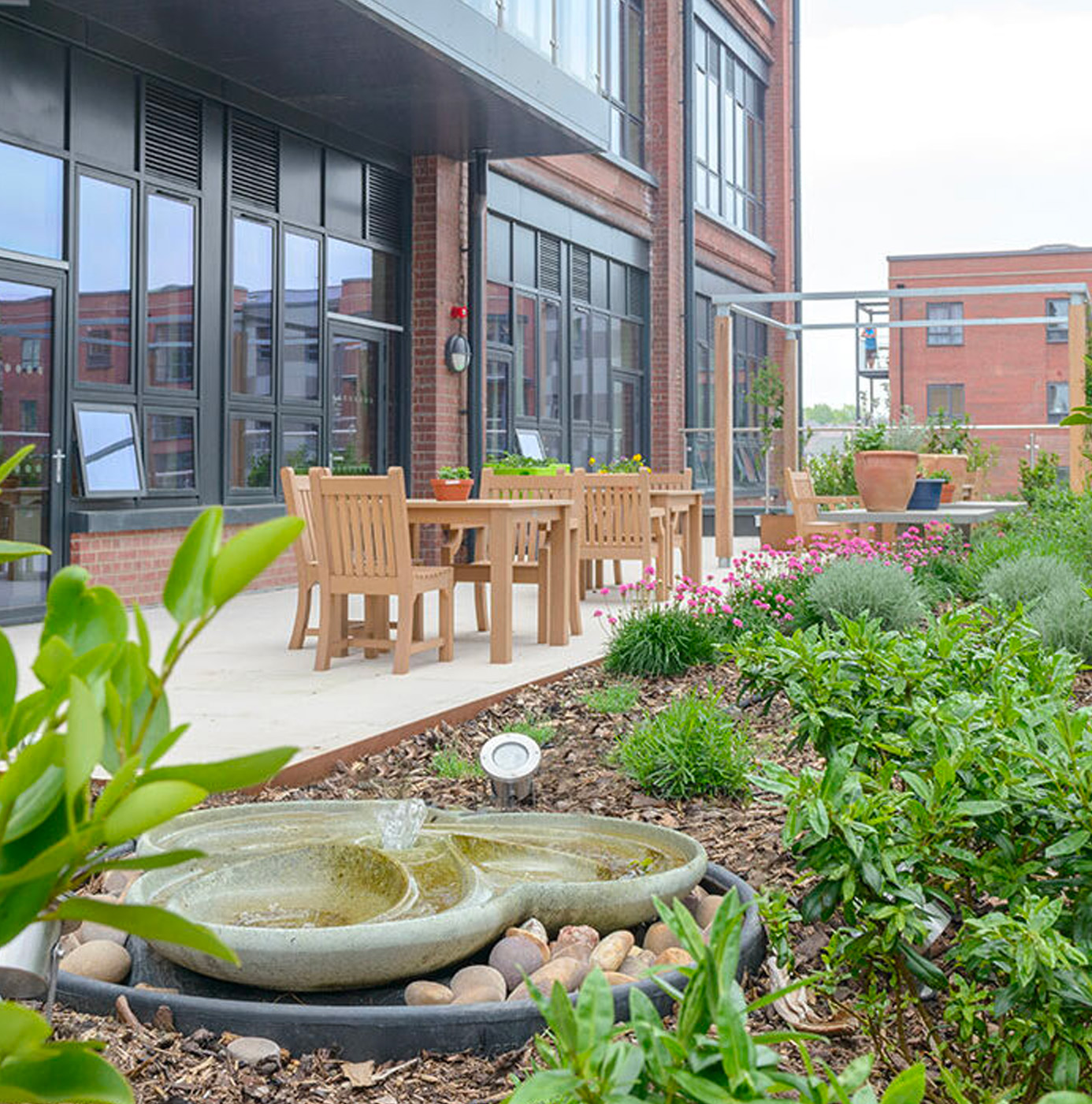 A planted rooftop terrace on a converted brick building with a stone water feature, mixed shrub and perennial borders, terracotta pots, and teak outdoor dining furniture.