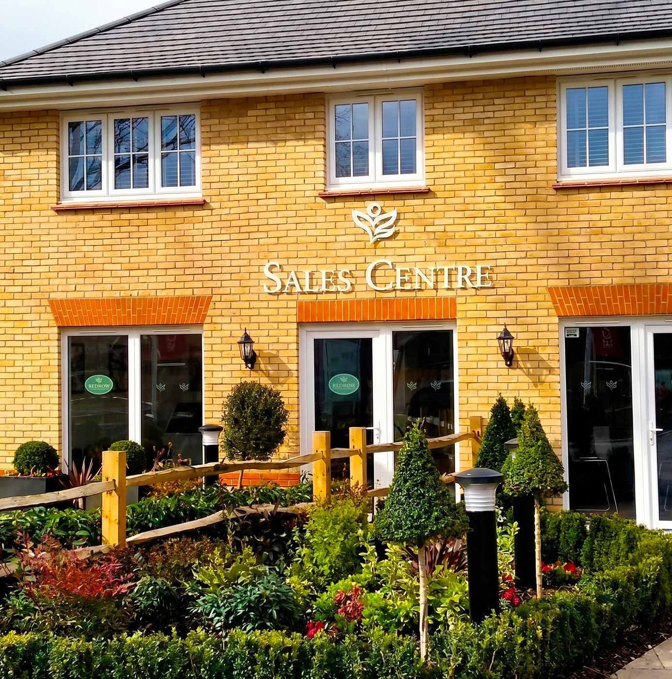 Soft landscaping scheme at a residential sales centre entrance featuring box topiary, ornamental grasses, and mixed shrub borders with timber post fencing.